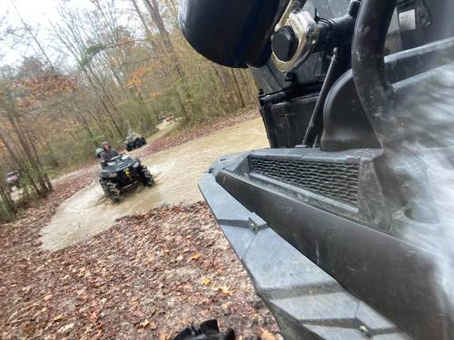 Two all-terrain vehicles navigate through a muddy area in a wooded setting during autumn.