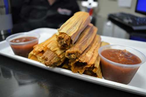 A plate of tamales stacked high, served with two small cups of red sauce on the side.
