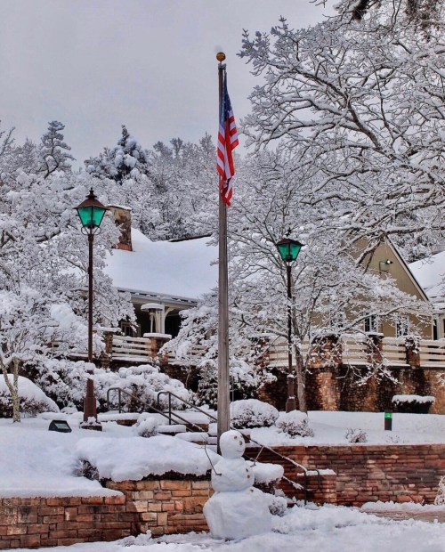 A snow-covered scene featuring a snowman, a flagpole with an American flag, and a cozy house in a winter landscape.