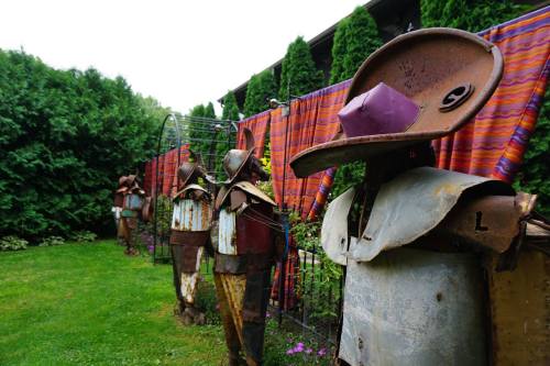 A garden scene featuring whimsical metal sculptures resembling scarecrows, set against colorful fabric and greenery.