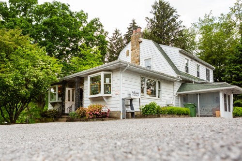 A charming white house with a sloped roof, surrounded by greenery and flowers, set on a gravel driveway.