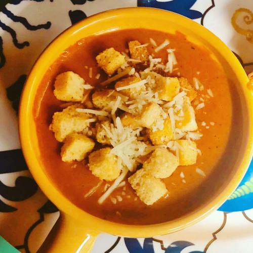 A bowl of tomato soup topped with croutons and shredded cheese, set on a colorful patterned table.