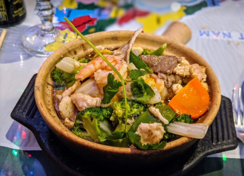 A clay pot filled with mixed stir-fried vegetables, shrimp, and beef, garnished with cilantro.