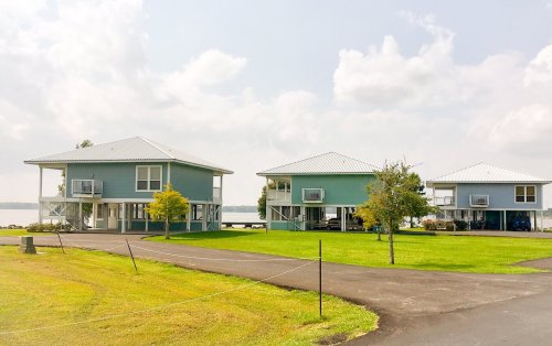 Three blue houses with porches on a grassy area by a lake, under a cloudy sky.