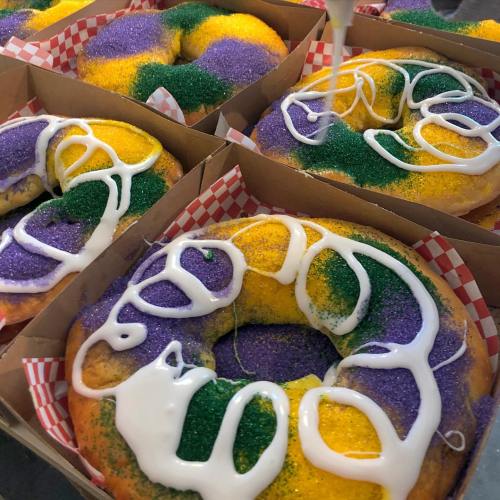 Colorful king cakes with purple, green, and yellow sprinkles, drizzled with white icing, arranged in cardboard boxes.