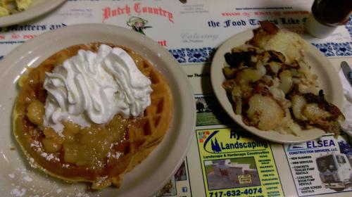 A plate with a waffle topped with whipped cream and apple topping, alongside a plate of crispy hash browns and sausage.