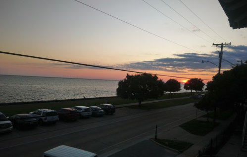 Sunset over a calm lake, with trees lining the shore and parked cars along the street in the foreground.