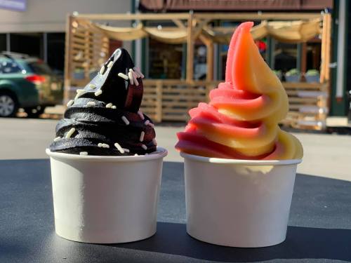 Two cups of soft serve ice cream: one black with sprinkles, the other pink and yellow swirled, set against a sunny background.