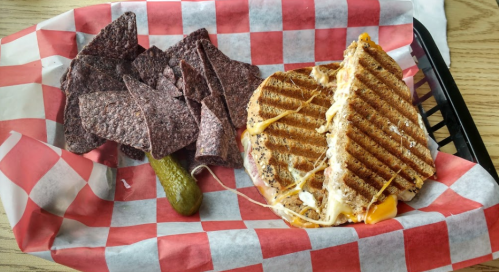 A grilled sandwich with melted cheese, a pickle, and blue corn tortilla chips on a red and white checkered tray.