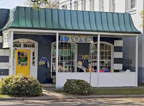 A charming toy store with a green roof, large windows, and colorful signage, surrounded by greenery.