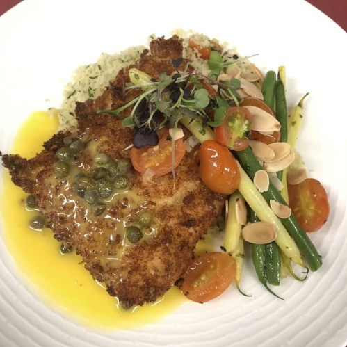 A plated dish featuring breaded fish with capers, served with green beans, cherry tomatoes, and a side of couscous.