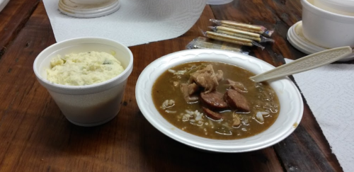 A bowl of stew with meat and sausage next to a cup of creamy mashed potatoes on a wooden table.