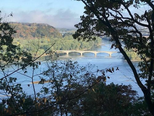 A scenic view of a river with a bridge, framed by trees and autumn foliage in the background.