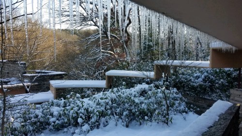Icicles hang from a roof above a snowy landscape with trees and shrubs, creating a serene winter scene.