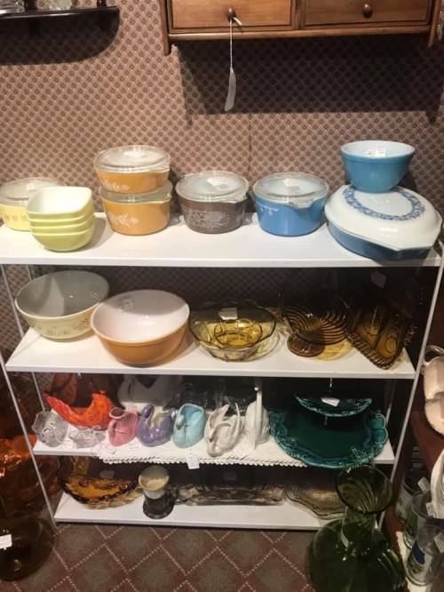 A display of vintage glassware and bowls in various colors on a white shelf against a patterned background.