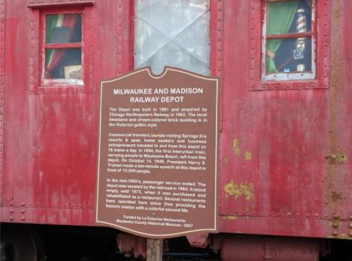 Sign about the Milwaukee and Madison Railway Depot, detailing its history and significance, next to a red train car.