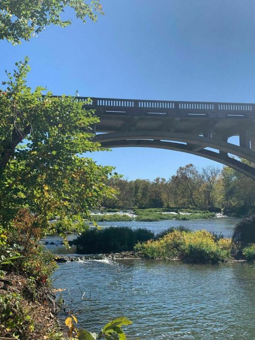 A bridge arches over a river surrounded by lush greenery and trees under a clear blue sky.