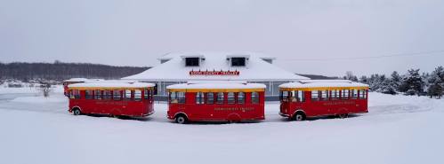 Two red trolleys with yellow roofs parked in front of a snowy building labeled "Door County Trolley." Winter landscape.