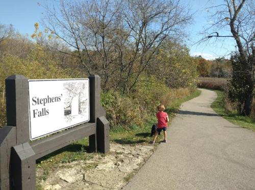 A child stands near a sign for Stephens' Falls, with a path and trees in the background on a sunny day.