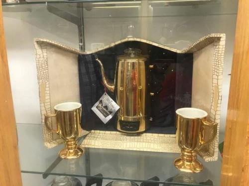 A gold-colored coffee pot with two matching cups displayed in a glass case, lined with a textured fabric.