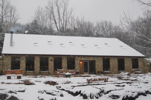A stone building covered in snow, surrounded by trees, with a warm light glowing from the entrance.