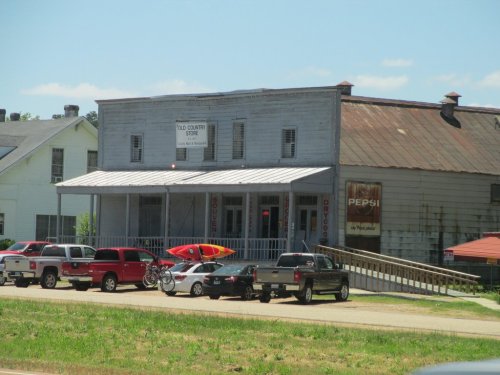 Old country store with a metal roof, porch, and a sign. A kayak and bicycles are outside, with parked cars nearby.