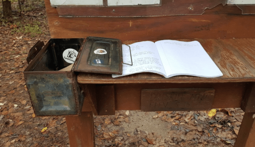 A wooden table in a forest with a metal box, a notebook, and a pen on top, surrounded by fallen leaves.