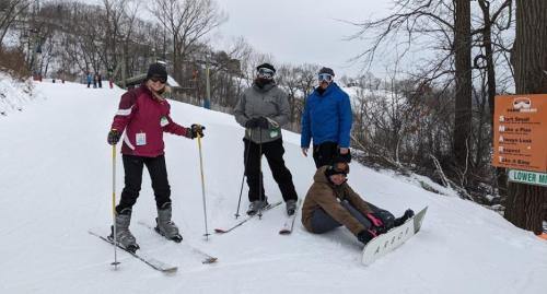 Four people on a snowy ski slope, two standing with skis, one sitting on a snowboard, and one in a blue jacket.