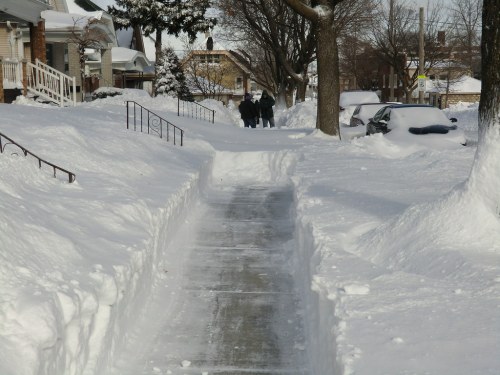 A snow-covered sidewalk with tall snowbanks on either side, two people walking in the distance.