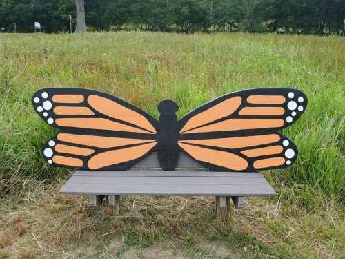 A wooden bench painted to resemble a butterfly, with orange wings and white spots, set in a grassy field.