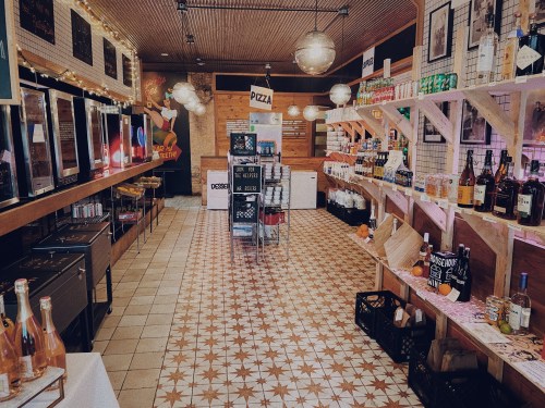 A cozy store interior with shelves of drinks, snacks, and pizza, featuring warm lighting and a patterned tile floor.