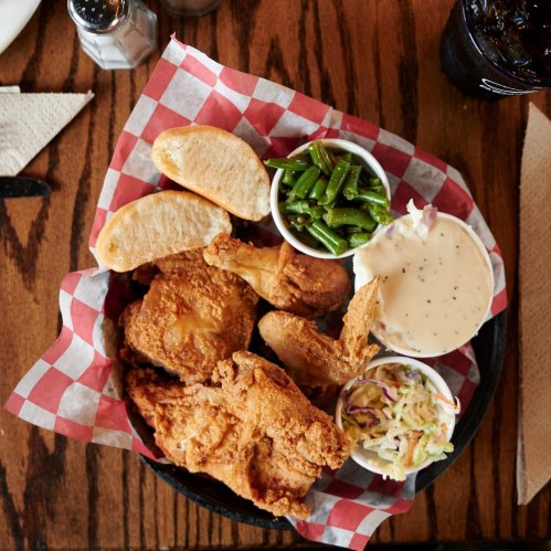 A basket of fried chicken with sides of green beans, coleslaw, and biscuits, served on a checkered paper.