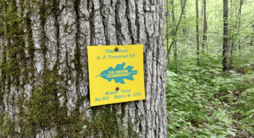 A yellow sign on a tree in a forest, indicating a nature preserve by The Nature Conservancy in Wisconsin.