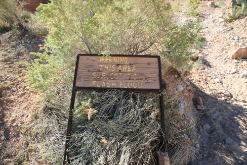 A weathered sign warns of potential flash floods in a desert area, surrounded by sparse vegetation.