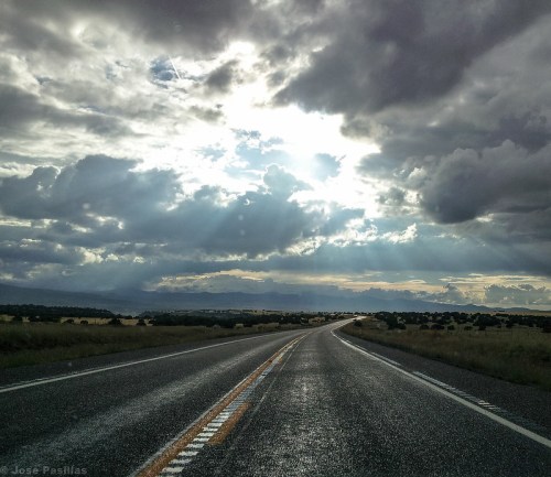 A winding road stretches into the distance under a dramatic sky with rays of sunlight breaking through the clouds.