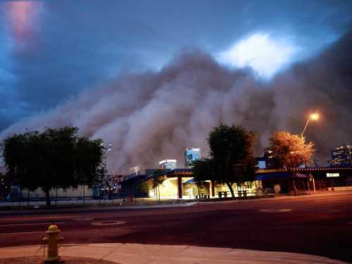 A dramatic cloud of dust looms over a cityscape at dusk, with trees and a streetlight in the foreground.