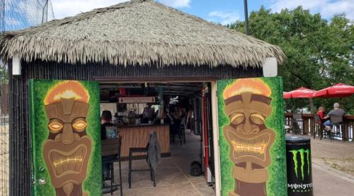 Tiki bar entrance with vibrant tiki carvings and a thatched roof, surrounded by outdoor seating and greenery.