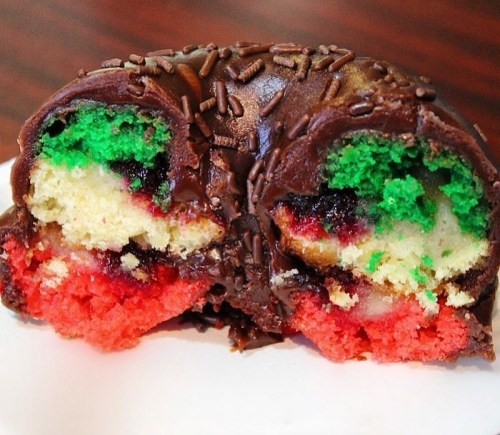 A cross-section of a colorful cake, featuring green, red, and yellow layers, coated in chocolate and topped with sprinkles.