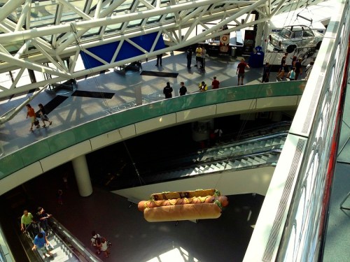 A large hot dog suspended from the ceiling in a busy indoor space with people and escalators below.