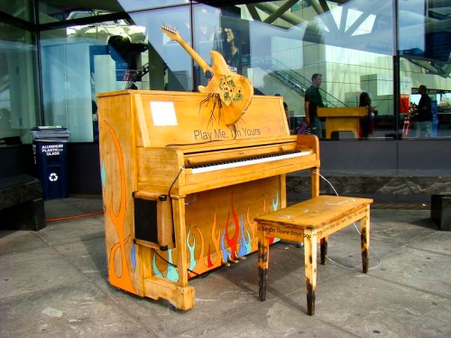 A colorful, painted piano with a guitar on top, set outside a building, accompanied by a matching stool.
