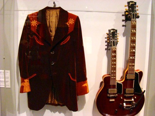 A vintage brown velvet jacket with red accents displayed alongside two electric guitars in a museum setting.