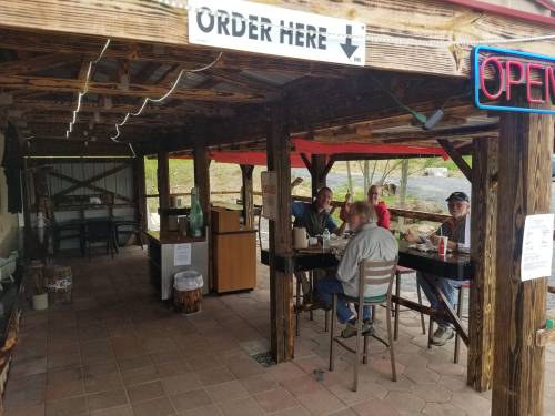 A rustic outdoor dining area with four people sitting at a table, a sign indicating "Order Here," and an "Open" neon sign.