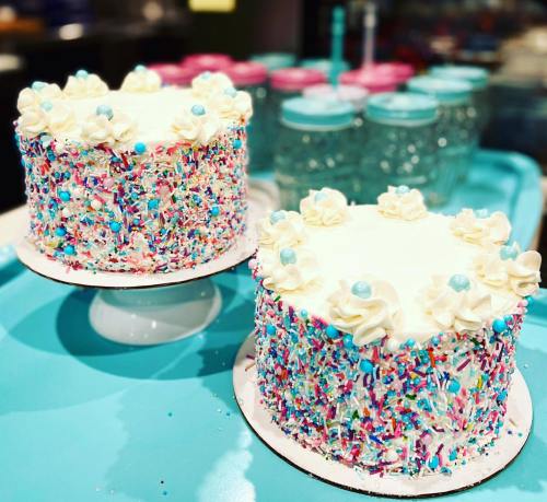 Two colorful cakes with white frosting, decorated with sprinkles and blue accents, on a turquoise surface.