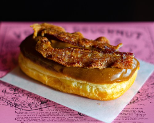 A glazed donut topped with a layer of peanut butter and crispy bacon strips, resting on a pink background.
