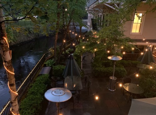 A cozy outdoor patio by a river, adorned with string lights and surrounded by greenery.