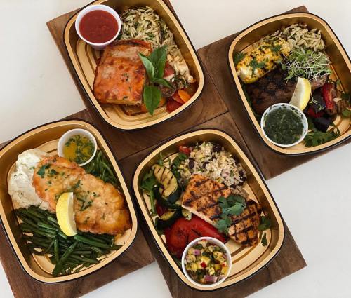 Four colorful meal boxes featuring grilled fish, rice, vegetables, and sauces, arranged on wooden trays.
