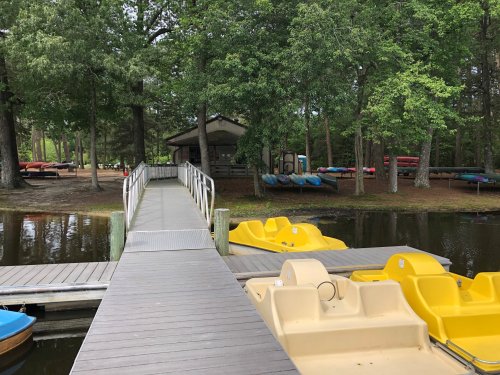 A wooden dock leads to a boathouse, with yellow paddle boats and kayaks lined up on the shore among green trees.
