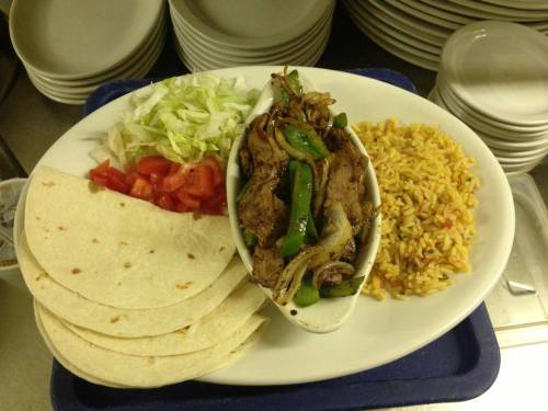 A platter with grilled meat, bell peppers, tortillas, lettuce, tomatoes, and seasoned rice, served on a white dish.