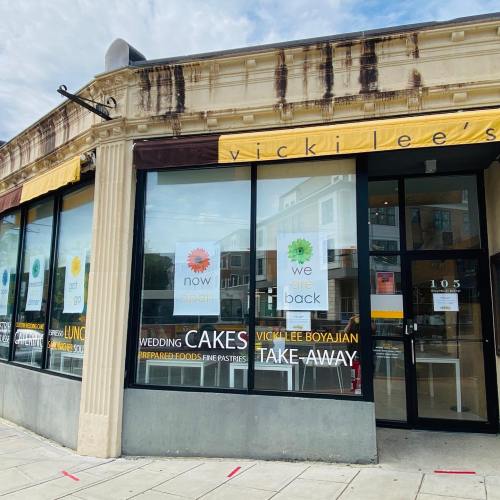 Exterior of Vicki Lee's bakery with large windows displaying signs for cakes, take-away, and reopening.