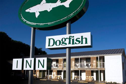 Sign for Dogfish Inn with a green logo and a view of the inn's exterior featuring balconies and a clear blue sky.